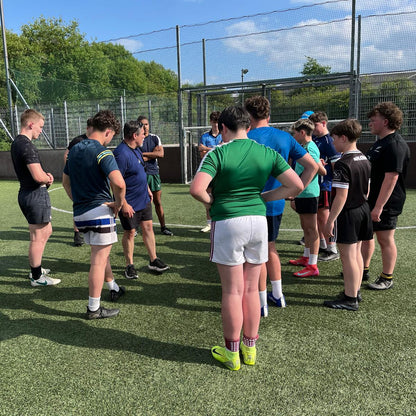 Group of young rugby players in a huddle on a sports field with green grass and a fence in the background.