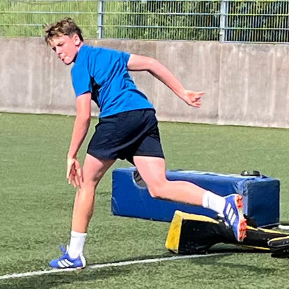 Person in blue shirt and shorts running on a sports field with a blue box and yellow item in the background.